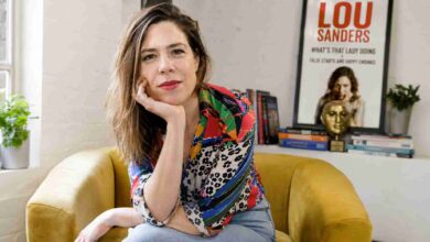 Lou Sanders sitting in a yellow armchair with her Taskmaster trophy and book poster in the background.