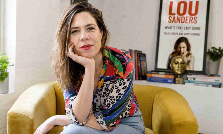 Lou Sanders sitting in a yellow armchair with her Taskmaster trophy and book poster in the background.