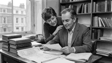 A vintage-style photograph of author Rayne Kruger sitting at a wooden desk reading a manuscript, with architectural blueprints spread out, as a younger Dame Prue Leith leans over his shoulder smiling.