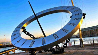A large steel equinoctial sundial suspended in mid-air by heavy black chains. This is the Grade II listed Timepiece sculpture created by Wendy Taylor, located at St Katharine Docks in London against a clear sky.