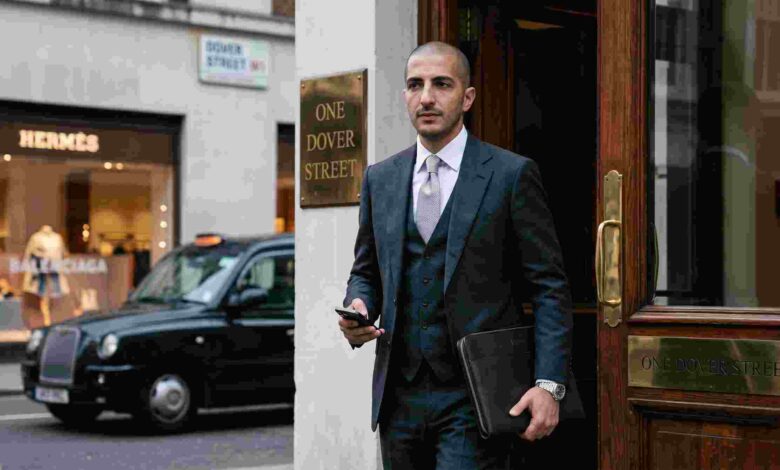 A candid photograph of Qatari businessman Wissam Al Mana in a grey three-piece suit walking out of the One Dover Street building in London. A Hermès store is visible in the background.